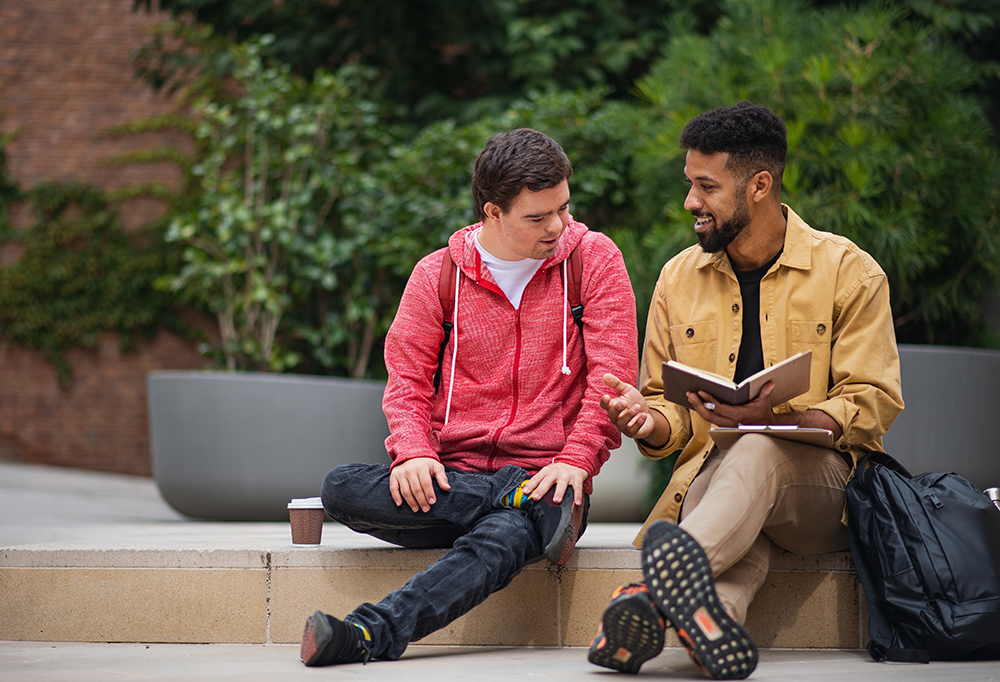 A happy young man with Down syndrome and mentoring friend sitting and talking.