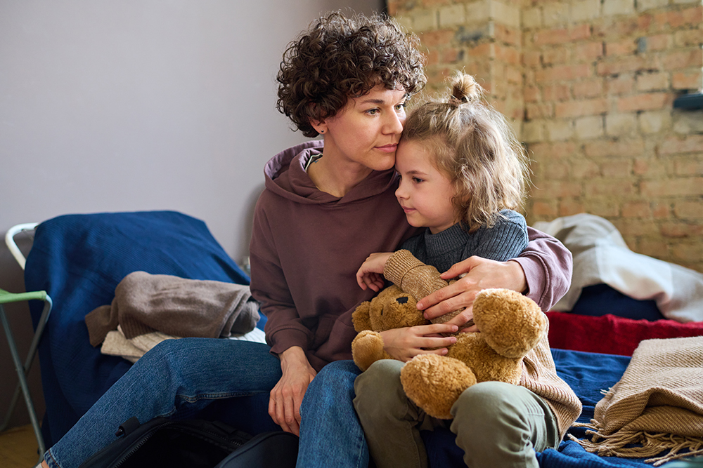 Young woman giving hug to her cute little son with brown soft teddybear while both sitting on sleeping pad at shelter