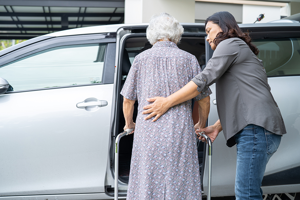 Caregiver supporting senior lady with walker into vehicle.